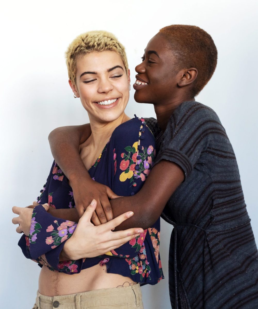 Photo of joyful women kissing and embracing togetherness, being of different of races, dressed in casual clothes.